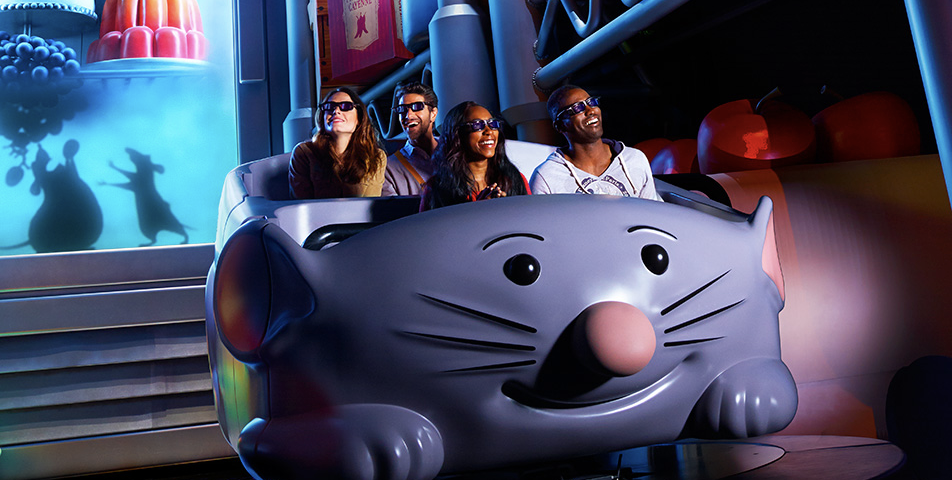 Quatre personnes portant des lunettes 3D montent dans une voiture grise en forme de souris dans l'attraction Ratatouille, un parc d'attractions coloré et animé qui a été inauguré le 10 juillet 2014.
