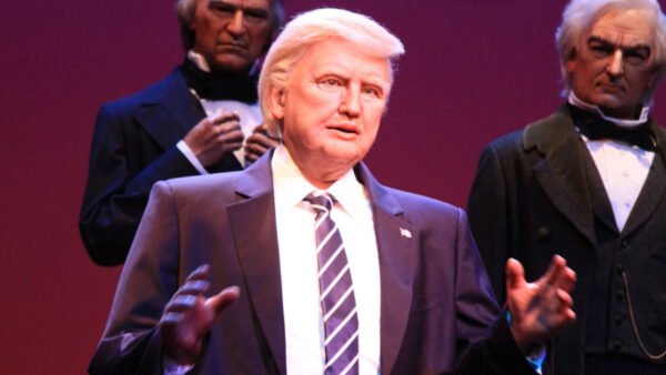 Un personnage de cire représentant Trump en costume et cravate rayée se tient devant deux autres personnages de cire en tenue de soirée sur un fond violet.