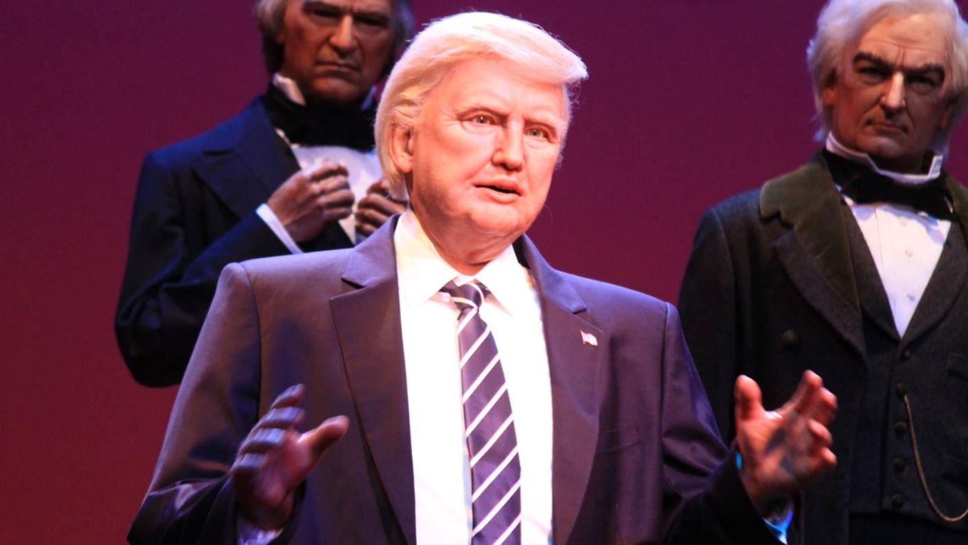 Un personnage de cire représentant Trump en costume et cravate rayée se tient devant deux autres personnages de cire en tenue de soirée sur un fond violet.