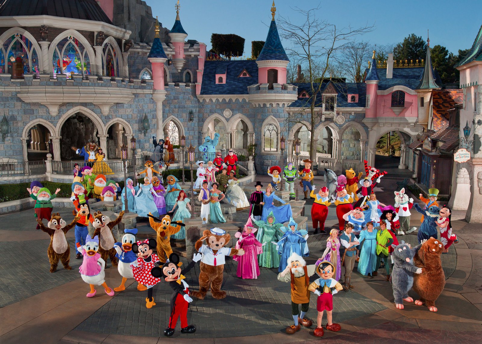 Un grand groupe de personnages Disneyland Paris en costume pose ensemble devant un château dans un parc à thème, capturant la magie des rencontres personnages Disneyland.