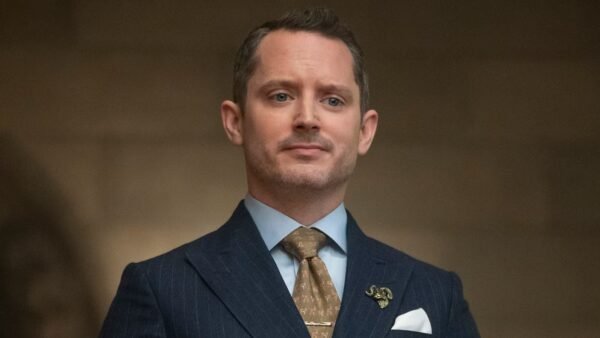 Un homme vêtu d'un costume à rayures sombres, d'une chemise bleu clair et d'une cravate à motifs se tient sur un fond neutre, regardant devant lui avec une expression neutre - ressemblant étrangement à Elijah Wood dans Paradise saison 3.