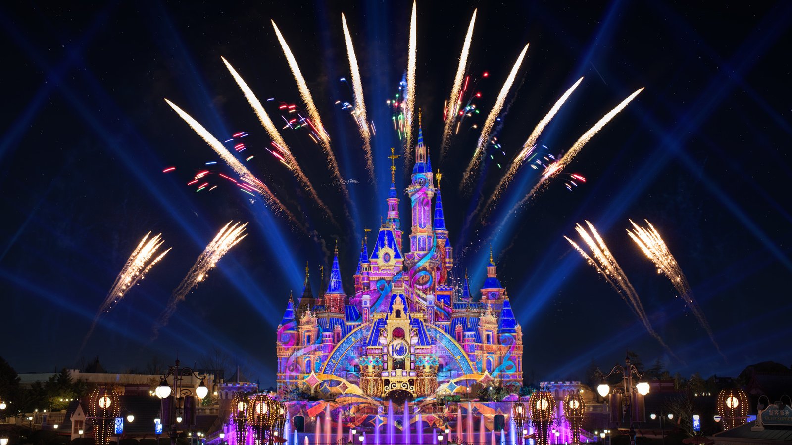 Le château de Shanghai Disneyland est illuminé de projections colorées la nuit, entouré de fontaines et de feux d'artifice qui s'élèvent dans le ciel.