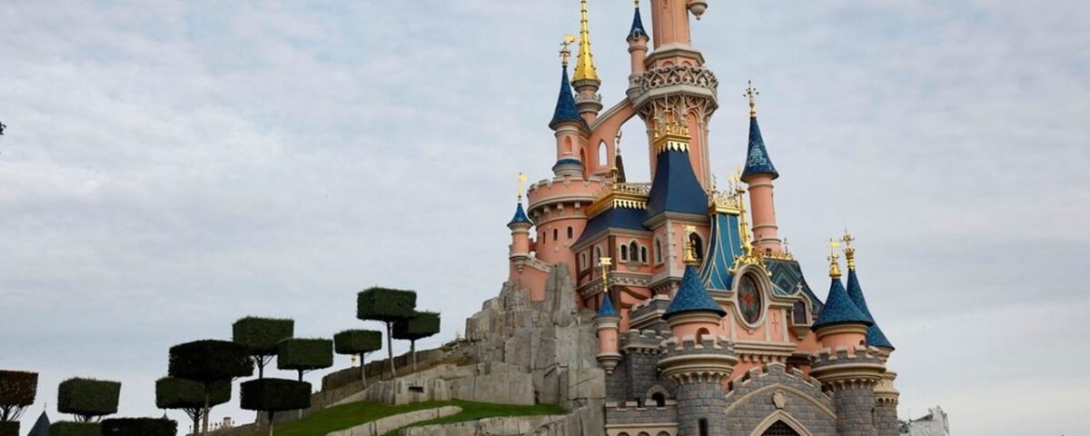 Un immense château de conte de fées, rose et bleu avec des touches dorées, se dresse sur un socle de pierre, entouré d'arbres soigneusement entretenus et de verdure, sous un ciel nuageux, évoquant la magie des forfaits Disneyland Paris.