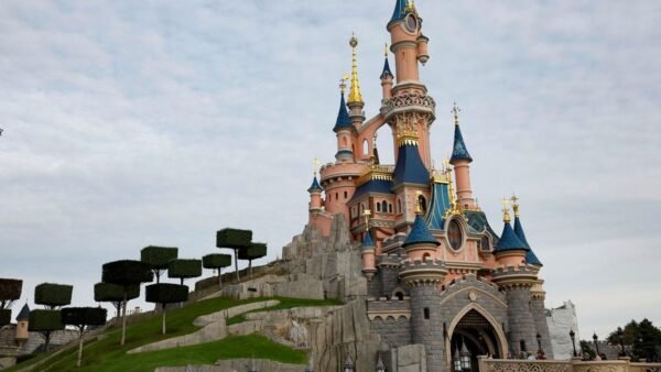 Un immense château de conte de fées, rose et bleu avec des touches dorées, se dresse sur un socle de pierre, entouré d'arbres soigneusement entretenus et de verdure, sous un ciel nuageux, évoquant la magie des forfaits Disneyland Paris.