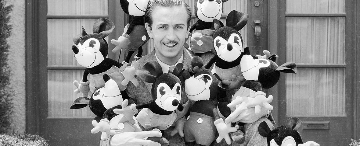 Un homme en costume est assis à l'extérieur, entouré de peluches Mickey Mouse, incarnant l'héritage Disney avec un clin d'œil ludique à l'esprit héritier Disney.
