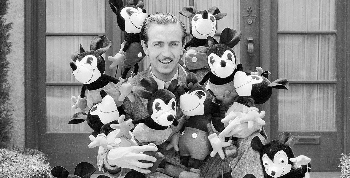 Un homme en costume est assis à l'extérieur, entouré de peluches Mickey Mouse, incarnant l'héritage Disney avec un clin d'œil ludique à l'esprit héritier Disney.