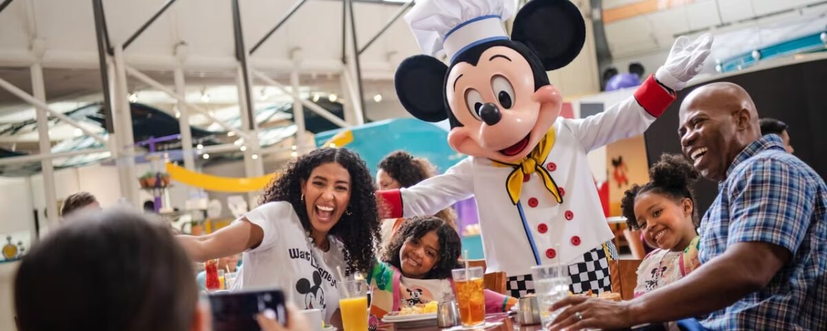 Une famille pose avec une personne déguisée en chef Mickey Mouse à la table d'un restaurant de Walt Disney World, souriant et dégustant des plats et des boissons à l'intérieur - un moment inoubliable rendu encore meilleur grâce aux réservations Disney World.