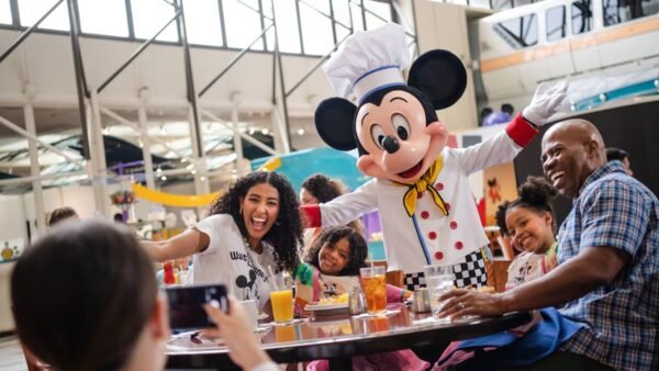 Une famille pose avec une personne déguisée en chef Mickey Mouse à la table d'un restaurant de Walt Disney World, souriant et dégustant des plats et des boissons à l'intérieur - un moment inoubliable rendu encore meilleur grâce aux réservations Disney World.