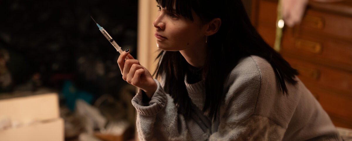 Une jeune femme aux cheveux noirs est assise à l'intérieur, tenant une seringue et regardant pensivement sur le côté, rappelant Emmy Rossum dans une scène d'un drame de Hulu.