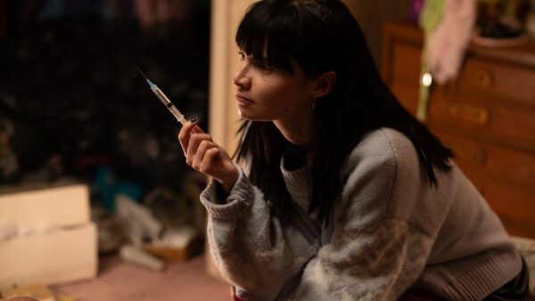 Une jeune femme aux cheveux noirs est assise à l'intérieur, tenant une seringue et regardant pensivement sur le côté, rappelant Emmy Rossum dans une scène d'un drame de Hulu.