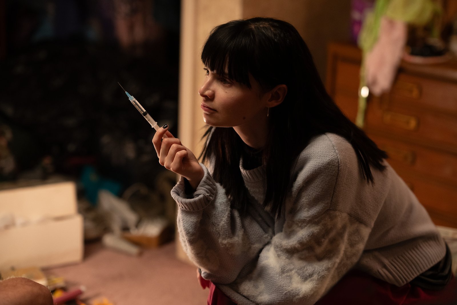 Une jeune femme aux cheveux noirs est assise à l'intérieur, tenant une seringue et regardant pensivement sur le côté, rappelant Emmy Rossum dans une scène d'un drame de Hulu.