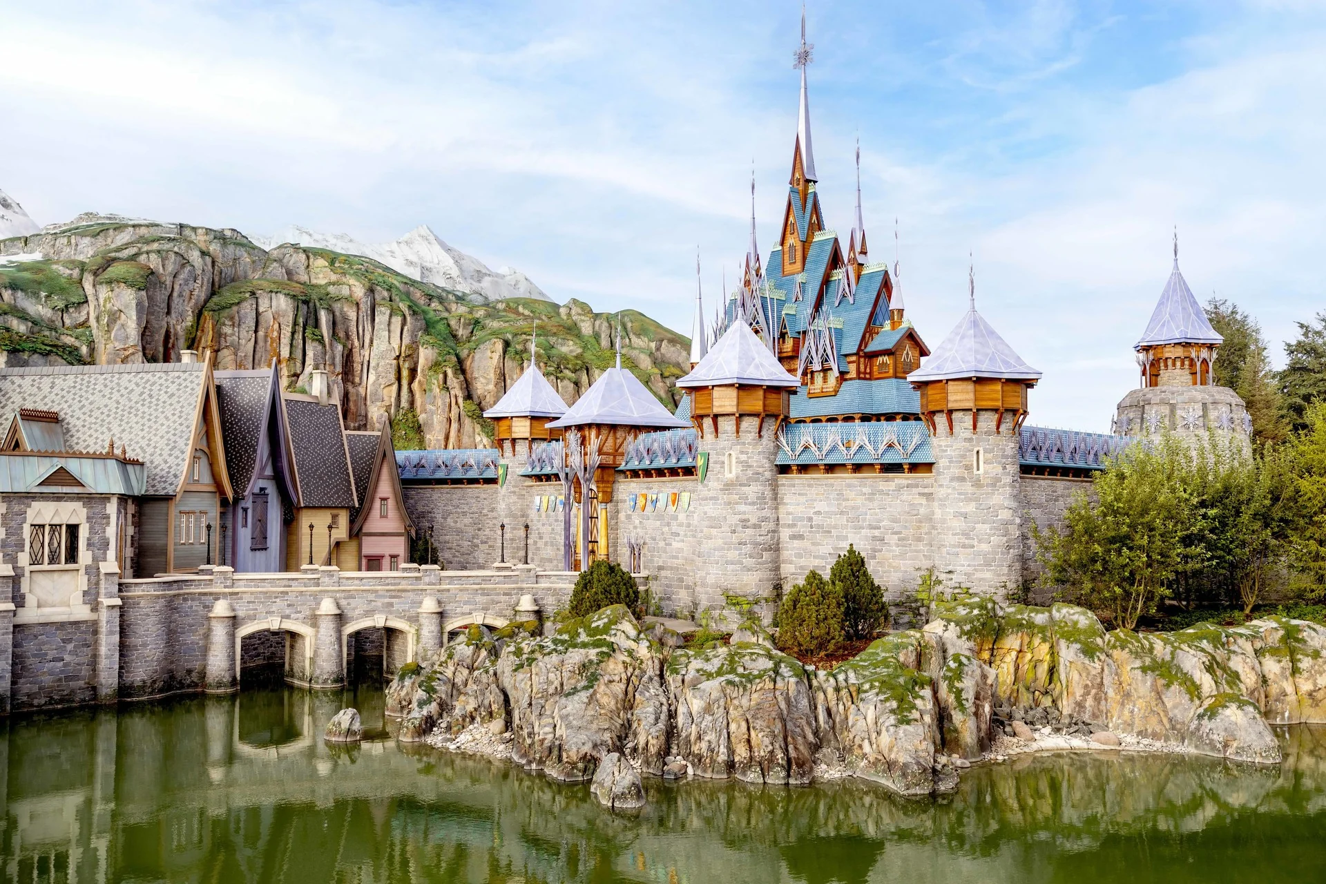 Un grand château de pierre orné aux toits bleus, inspiré du Château d'Arendelle, trône au bord d'un étang, entouré d'un paysage rocheux et de montagnes lointaines sous un ciel partiellement nuageux - évoquant la magie de Walt Disney Imagineering à Disneyland Paris.