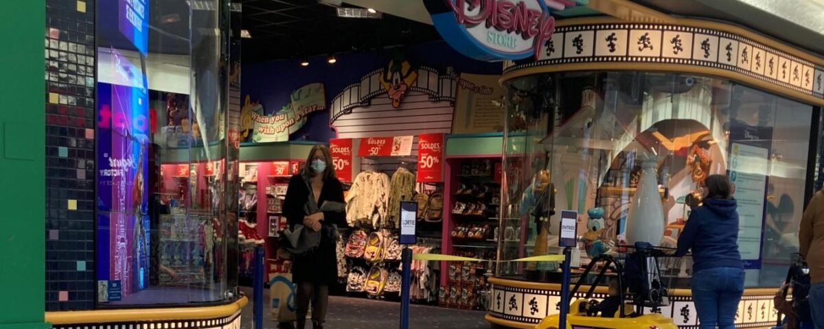 Un Disney Store dans un centre commercial avec des présentoirs colorés, des panneaux de vente et des clients qui cherchent des souvenirs Disney à l'intérieur et à l'extérieur, entourés d'éléments culturels dynamiques et de produits classiques de Disney.
