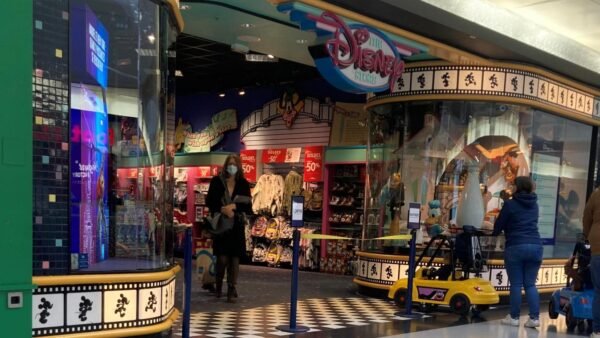 Un Disney Store dans un centre commercial avec des présentoirs colorés, des panneaux de vente et des clients qui cherchent des souvenirs Disney à l'intérieur et à l'extérieur, entourés d'éléments culturels dynamiques et de produits classiques de Disney.