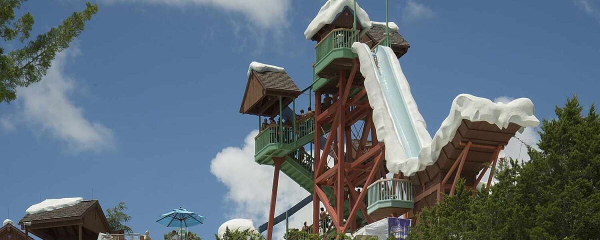 Une grande tour de toboggans aquatiques décorée sur le thème de la neige à Blizzard Beach, avec des gens qui attendent à ses pieds, sous un ciel partiellement nuageux et au milieu d'un cadre verdoyant.