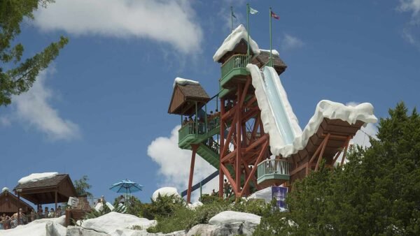 Une grande tour de toboggans aquatiques décorée sur le thème de la neige à Blizzard Beach, avec des gens qui attendent à ses pieds, sous un ciel partiellement nuageux et au milieu d'un cadre verdoyant.