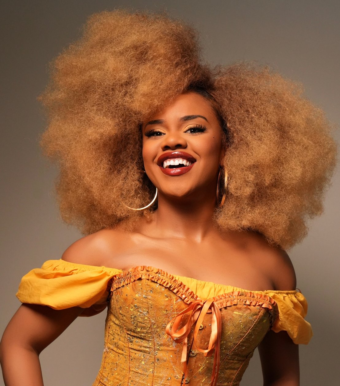 Une femme aux cheveux bouclés volumineux porte une robe jaune décolletée et de grandes boucles d'oreilles, souriant sur un fond uni - son look est le reflet du style vibrant des Descendants.