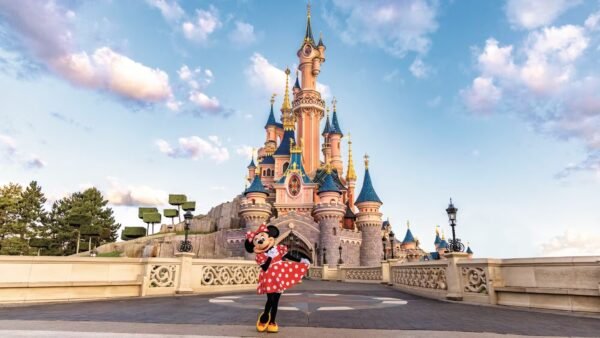 Une personne déguisée en Minnie Mouse pose devant un château de conte de fées rose et bleu, sous un ciel bleu parsemé de nuages — l'image parfaite pour annoncer un séjour magique à Disneyland Paris et trouver des idées de photos inoubliables.