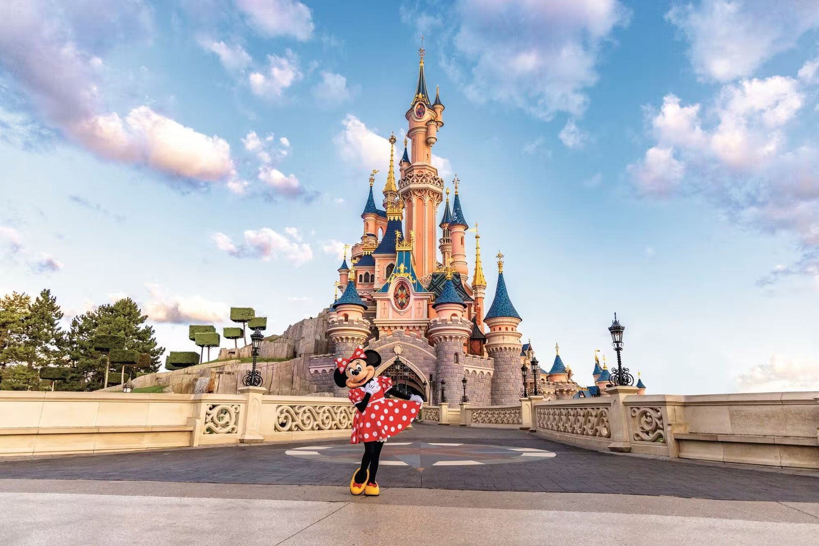 Une personne déguisée en Minnie Mouse pose devant un château de conte de fées rose et bleu, sous un ciel bleu parsemé de nuages — l'image parfaite pour annoncer un séjour magique à Disneyland Paris et trouver des idées de photos inoubliables.