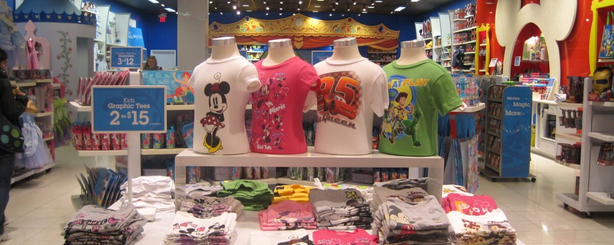Présentation de T-shirts pour enfants avec des produits dérivés Disney représentant des personnages bien-aimés sur des mannequins et des piles pliées dans un magasin de détail bien éclairé, présentant de véritables exemples de succès Disney.
