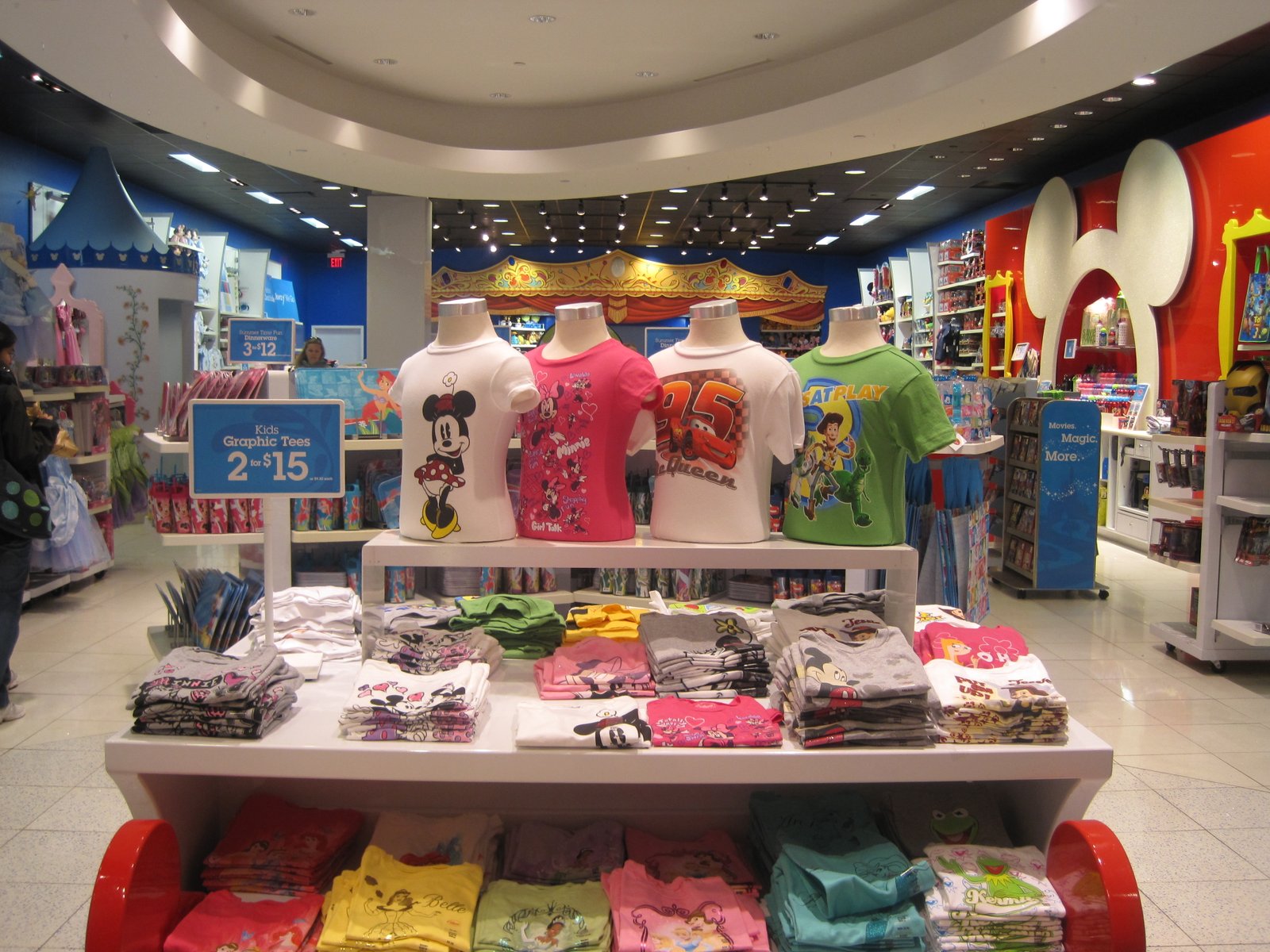 Présentation de T-shirts pour enfants avec des produits dérivés Disney représentant des personnages bien-aimés sur des mannequins et des piles pliées dans un magasin de détail bien éclairé, présentant de véritables exemples de succès Disney.