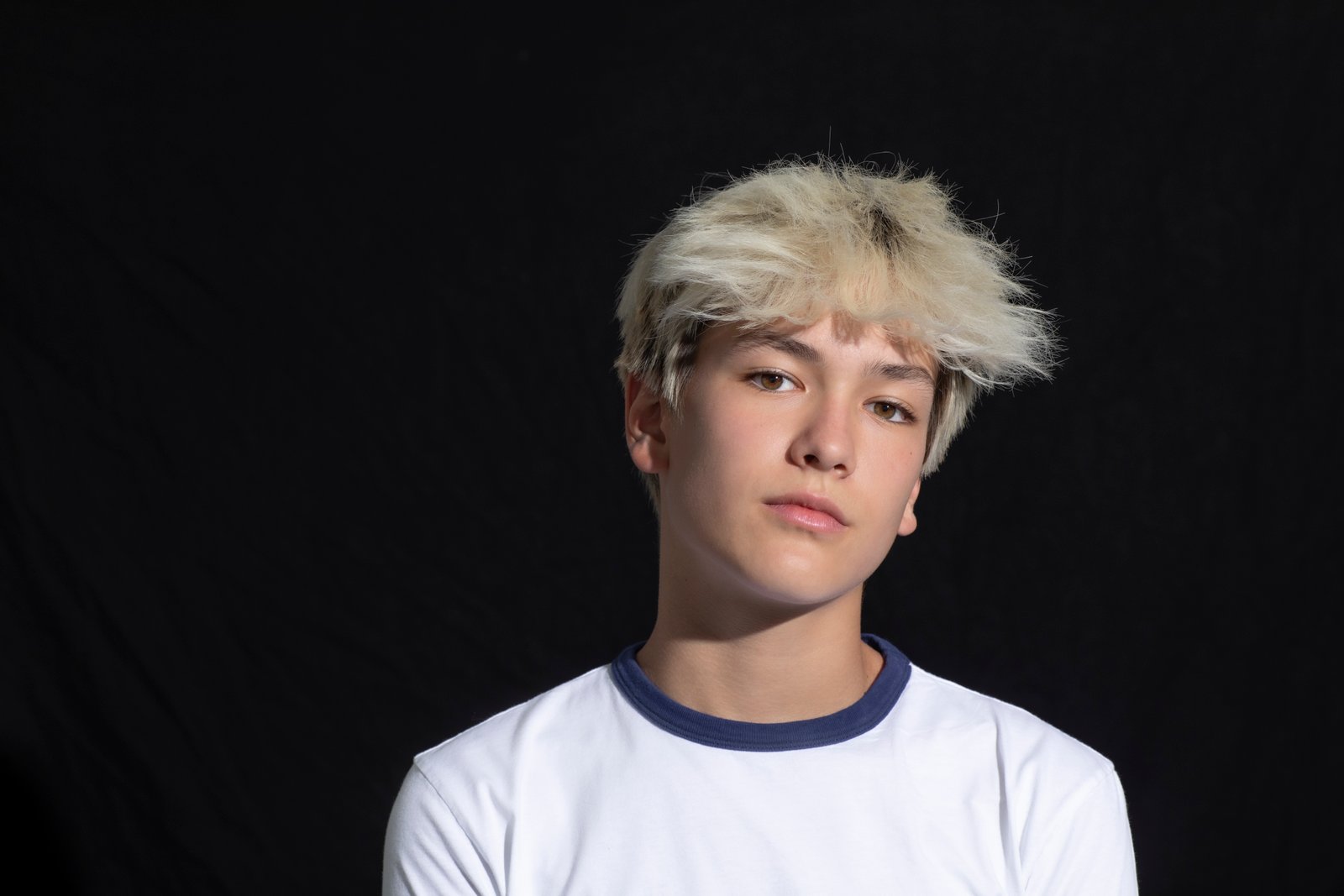 Adolescent aux cheveux blonds courts et désordonnés portant un t-shirt blanc avec des bordures bleues, posant devant un fond noir uni - canalisant l'esprit de Stars De Descendants.