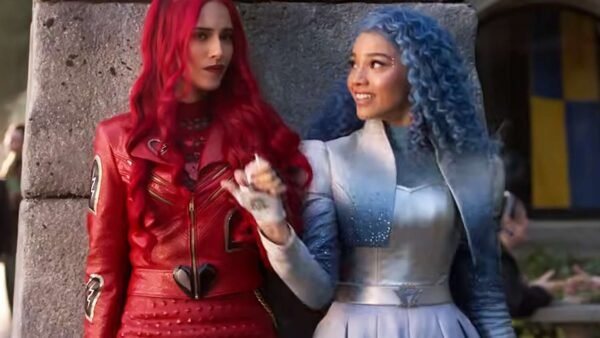 Deux femmes se tiennent côte à côte ; l'une a de longs cheveux roux et une tenue de cuir rouge, tandis que l'autre, dans un costume bleu argenté et des cheveux bleus bouclés, sourit - ce qui rappelle Descendants de Disney Channel ou La Malédiction Du Pays Des Merveilles.