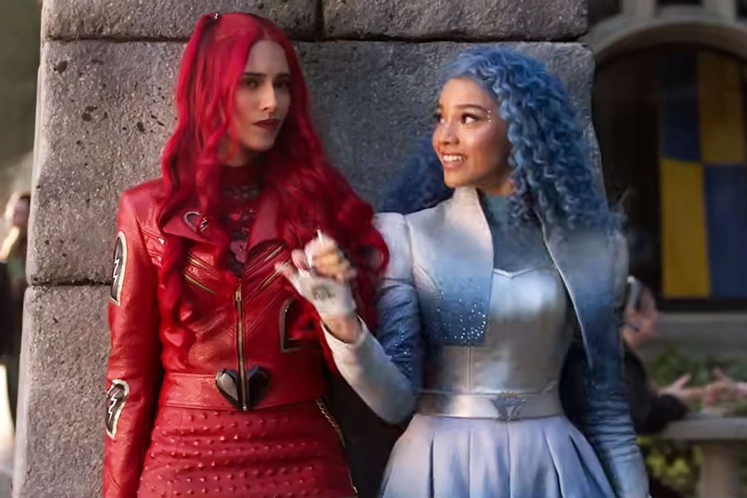Deux femmes se tiennent côte à côte ; l'une a de longs cheveux roux et une tenue de cuir rouge, tandis que l'autre, dans un costume bleu argenté et des cheveux bleus bouclés, sourit - ce qui rappelle Descendants de Disney Channel ou La Malédiction Du Pays Des Merveilles.