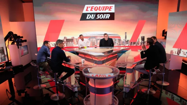 Un groupe de six personnes est assis autour d'une table en verre sur le plateau de "L'Équipe du Soir", une émission de télévision sportive diffusée sur La Chaîne L'Équipe, avec des lumières de studio et des caméras visibles à l'arrière-plan.