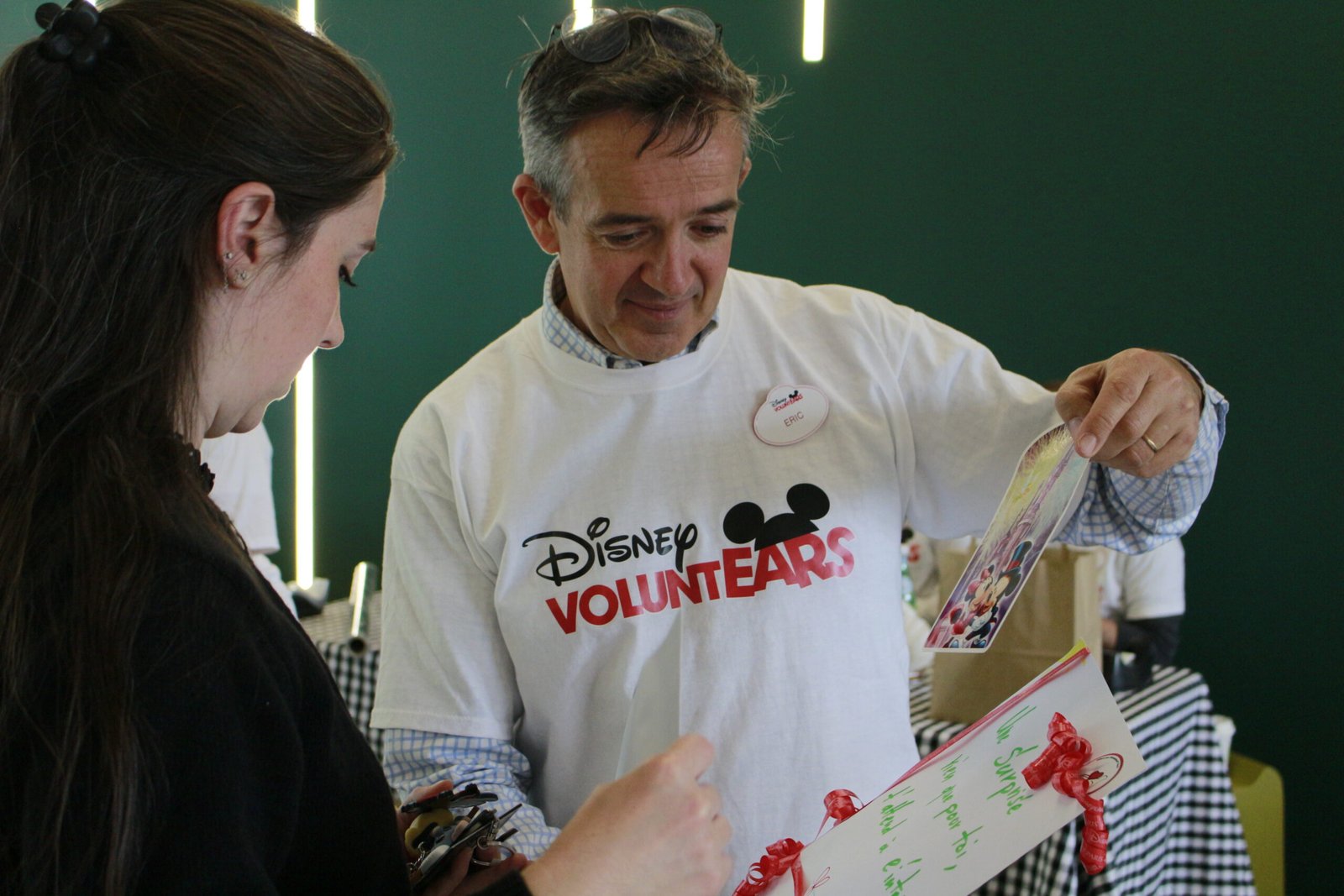 Deux personnes regardent une carte décorée ; l'une d'elles porte un t-shirt « Disney VoluntEARS » et un badge nominatif, et participe à une activité organisée dans le cadre de la Semaine des bénévoles Disney à Disneyland Paris.