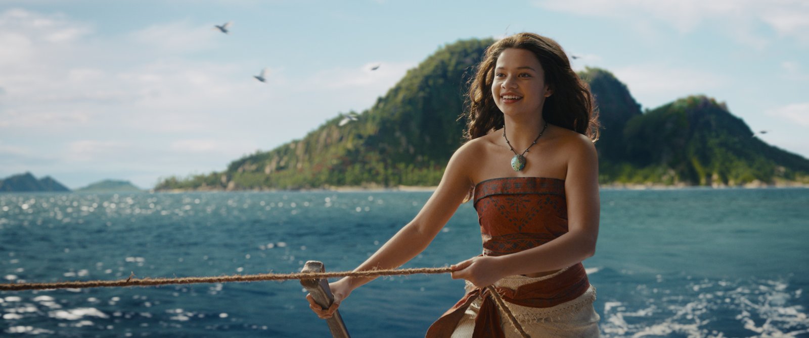 Une jeune femme vêtue d'une robe à motifs se tient debout sur un bateau, une corde à la main, avec l'océan et une île verdoyante et montagneuse en arrière-plan — tout comme Vaiana, l'héroïne courageuse et aventureuse de Disney.