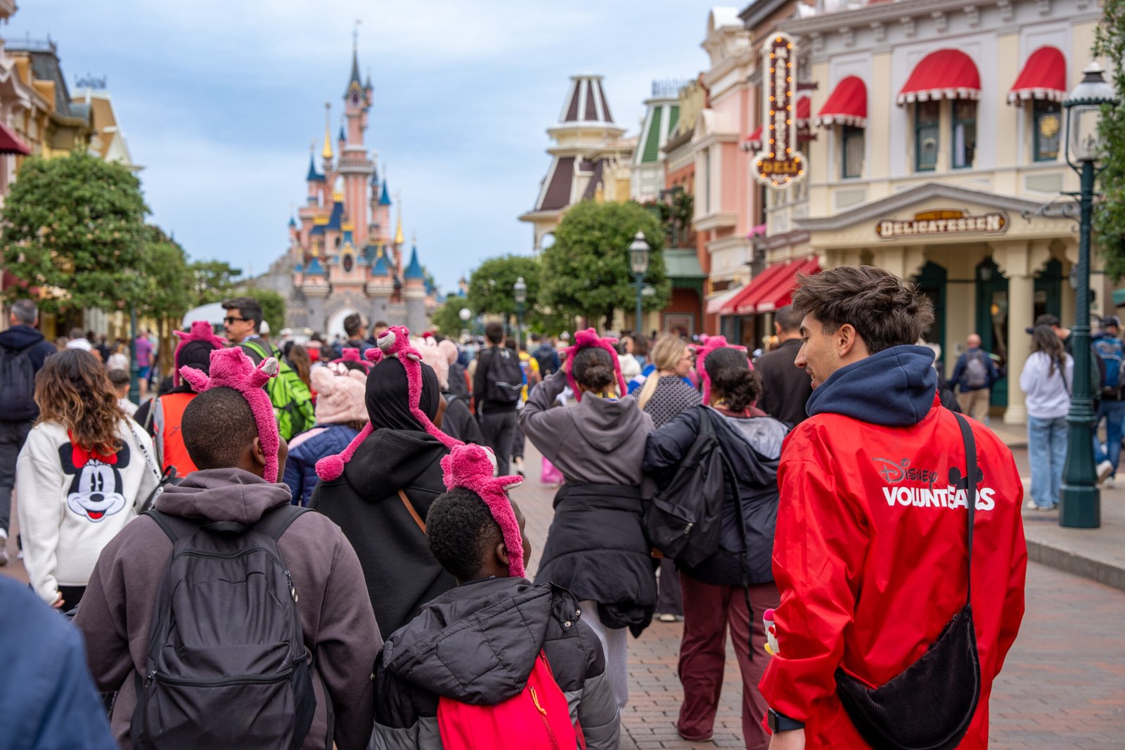 Un groupe de personnes, dont certaines portent des oreilles roses de Minnie Mouse, se dirige vers un château à Disneyland Paris, accompagné d'une personne vêtue d'une veste rouge Disney Voluntears.