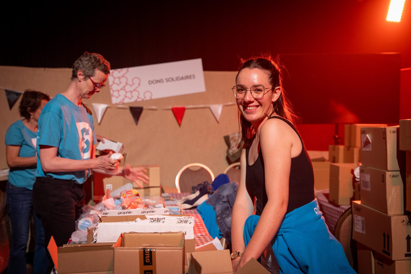Une femme sourit à la caméra tout en triant des dons sur une table dans le cadre du programme Disney Voluntears ; d'autres bénévoles s'affairent à proximité avec des cartons et du matériel, sous un éclairage chaleureux.