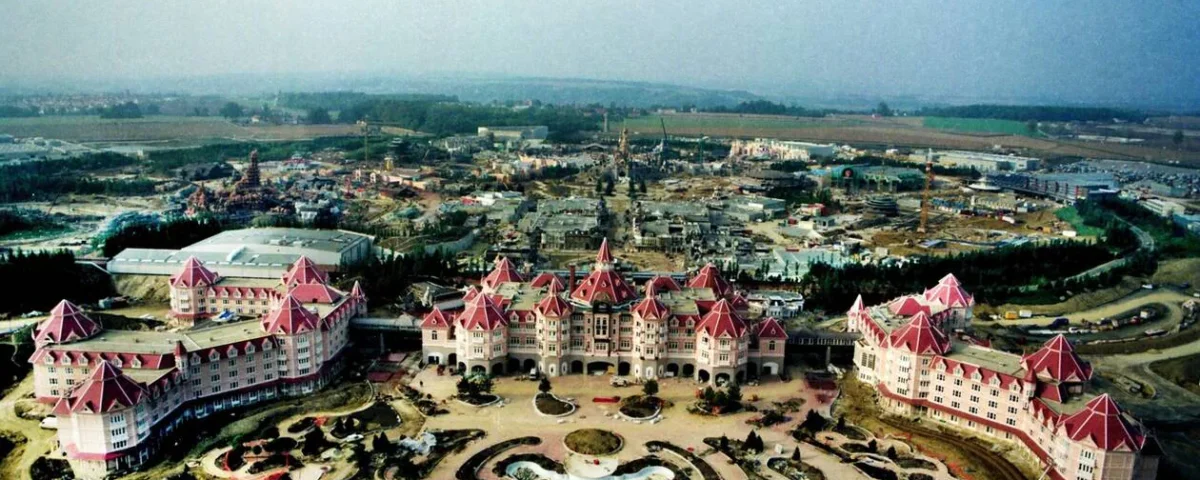 Vue aérienne d'un grand hôtel au toit rose avec des jardins paysagers devant, entouré de chantiers et de champs, rappelant les zones thématiques de Disneyland Paris pour une expérience visiteur unique.