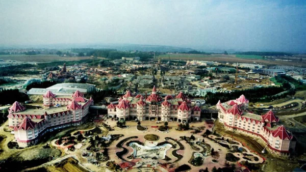 Vue aérienne d'un grand hôtel au toit rose avec des jardins paysagers devant, entouré de chantiers et de champs, rappelant les zones thématiques de Disneyland Paris pour une expérience visiteur unique.