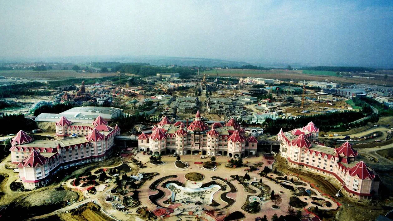 Vue aérienne d'un grand hôtel au toit rose avec des jardins paysagers devant, entouré de chantiers et de champs, rappelant les zones thématiques de Disneyland Paris pour une expérience visiteur unique.
