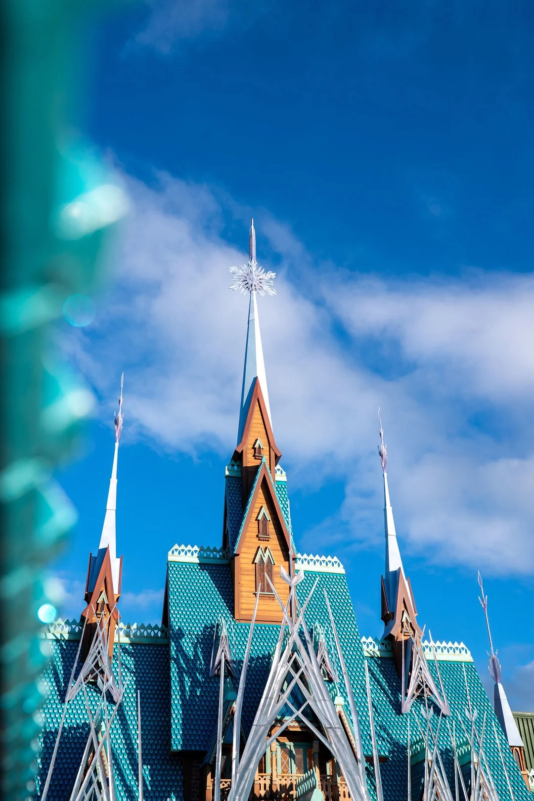 Un bâtiment avec des toits pointus en tuiles bleues et un ornement en forme de flocon de neige au sommet, inspiré du château d'Arendelle, sur fond de ciel bleu vif avec quelques nuages.