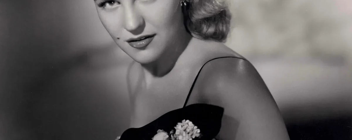 Portrait en noir et blanc d'une femme aux cheveux blonds ondulés, vêtue d'une robe sombre ornée d'appliqués floraux, qui regarde l'objectif avec une expression neutre — évoquant l'élégance de Peggy Lee dans *La Belle et le Clochard* et les chansons classiques de Disney.