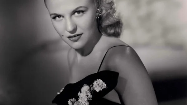 Portrait en noir et blanc d'une femme aux cheveux blonds ondulés, vêtue d'une robe sombre ornée d'appliqués floraux, qui regarde l'objectif avec une expression neutre — évoquant l'élégance de Peggy Lee dans *La Belle et le Clochard* et les chansons classiques de Disney.