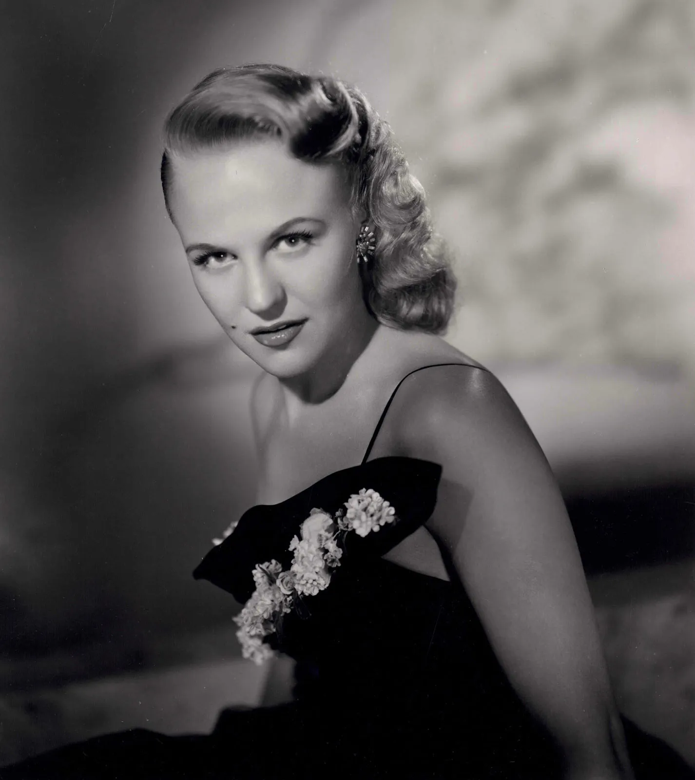 Portrait en noir et blanc d'une femme aux cheveux blonds ondulés, vêtue d'une robe sombre ornée d'appliqués floraux, qui regarde l'objectif avec une expression neutre — évoquant l'élégance de Peggy Lee dans *La Belle et le Clochard* et les chansons classiques de Disney.