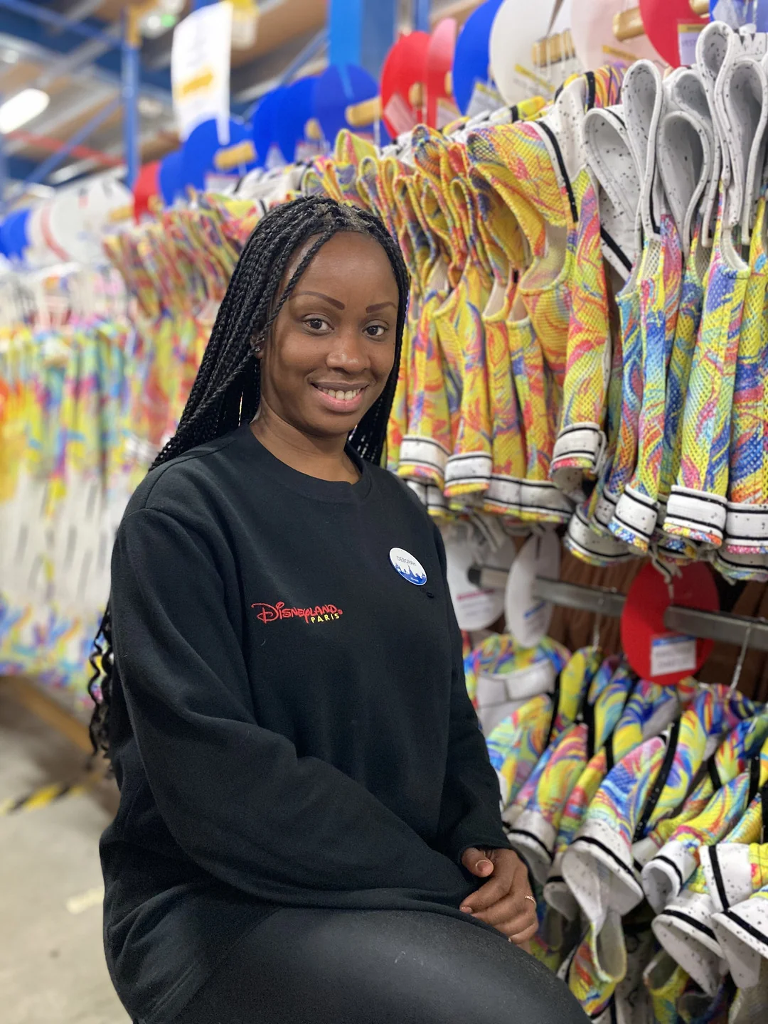 Une femme vêtue d'un sweat-shirt noir sur le thème de Disneyland Paris sourit en posant devant des rayonnages de chaussures à motifs colorés dans un entrepôt ou une usine.