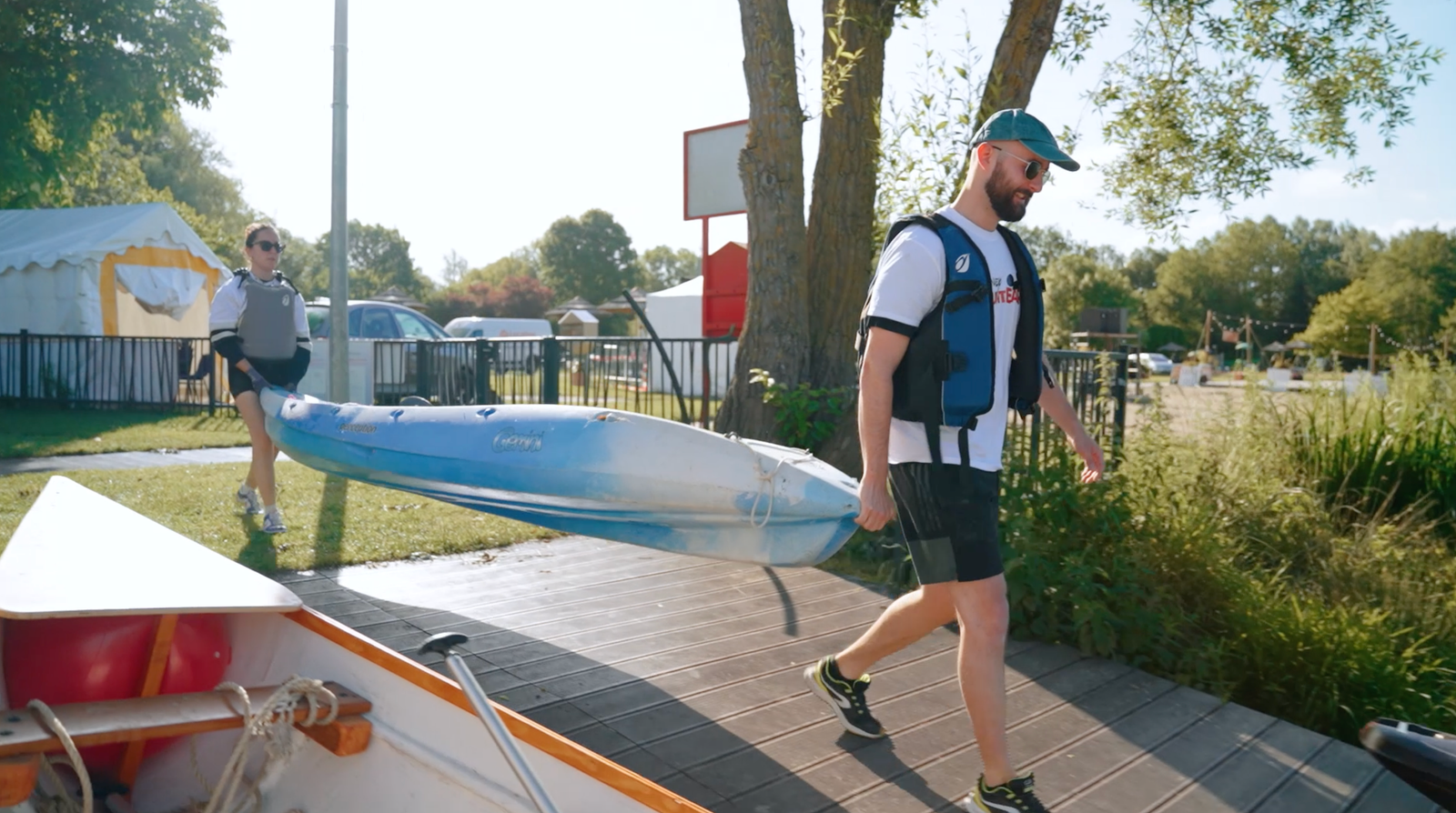 Par une journée ensoleillée, deux personnes vêtues de gilets de sauvetage transportent un kayak bleu et blanc le long d'un quai en bois, près d'arbres et de voitures garées, illustrant ainsi l'esprit de volontariat inspiré par Disney Voluntears.
