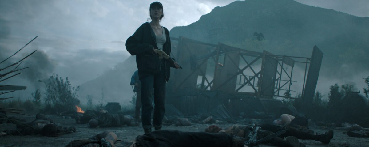 Une femme tenant un fusil se tient au milieu de cadavres sur un champ de bataille enfumé et jonché de débris, évoquant un thriller épique de Ridley Scott, avec une structure endommagée et des montagnes se profilant à l'arrière-plan.