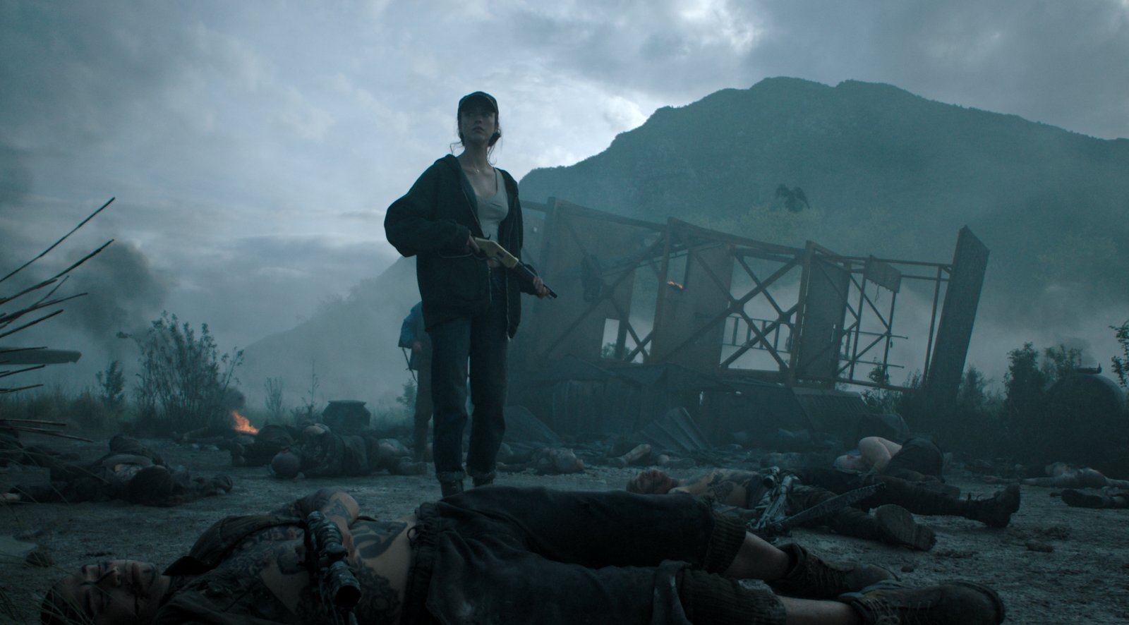 Une femme tenant un fusil se tient au milieu de cadavres sur un champ de bataille enfumé et jonché de débris, évoquant un thriller épique de Ridley Scott, avec une structure endommagée et des montagnes se profilant à l'arrière-plan.
