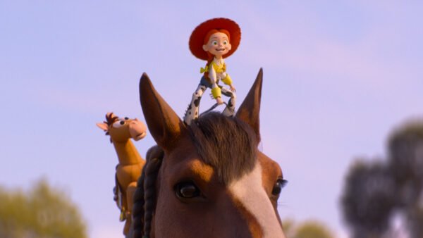 Une poupée avec un chapeau rouge et des vêtements jaunes est assise en souriant sur la tête d'un cheval, avec un cheval jouet à proximité - comme dans une scène de Toy Story 5. L'arrière-plan est un ciel clair avec des arbres flous.