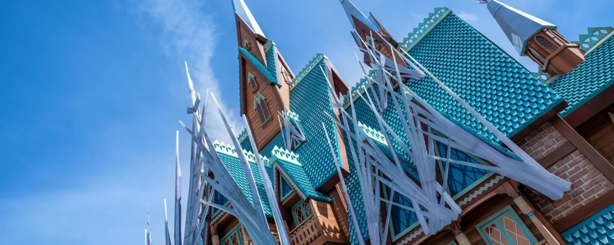 Un bâtiment aux toits de tuiles bleues, aux balcons en bois et aux hautes flèches pointues, inspiré du Château d'Arendelle, présente des décorations blanches ressemblant à des glaçons sous un ciel bleu clair.