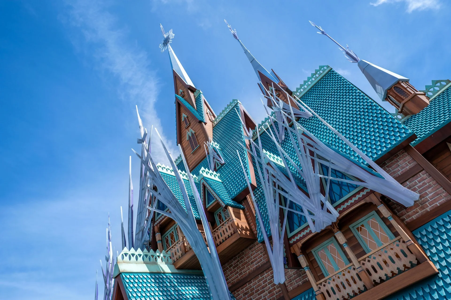 Un bâtiment aux toits de tuiles bleues, aux balcons en bois et aux hautes flèches pointues, inspiré du Château d'Arendelle, présente des décorations blanches ressemblant à des glaçons sous un ciel bleu clair.