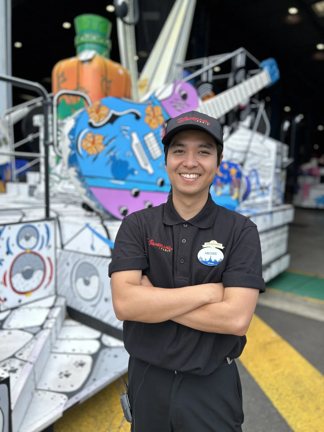 Un Cast Member en uniforme et casquette noirs sourit, les bras croisés, devant un char coloré de la parade, orné d'une grande guitare et de motifs de haut-parleurs, capturant l'unique Magie Disney à Disneyland Paris.