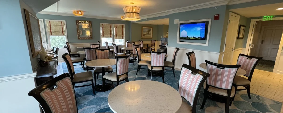Un salon bien éclairé avec des tables rondes, des chaises rembourrées à rayures, des murs bleus, des œuvres d'art et une télévision murale - parfait pour se détendre ou recevoir l'assistance d'un concierge Aux Invités inspiré par l'hospitalité de Walt Disney World.