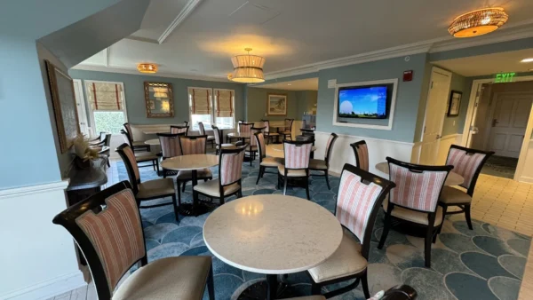 Un salon bien éclairé avec des tables rondes, des chaises rembourrées à rayures, des murs bleus, des œuvres d'art et une télévision murale - parfait pour se détendre ou recevoir l'assistance d'un concierge Aux Invités inspiré par l'hospitalité de Walt Disney World.